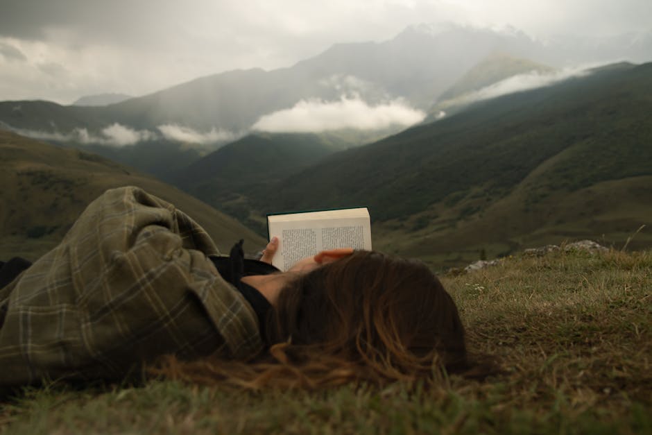 A person reads peacefully on a grassy hill with a stunning mountain backdrop and cloudy sky.